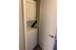 A stacked washer and dryer unit located in a narrow laundry space, positioned next to a closed white door. The floor features light wood-like laminate, and there's a small vent on the wall below the door. The walls are painted a neutral color, creating a compact and functional laundry area.