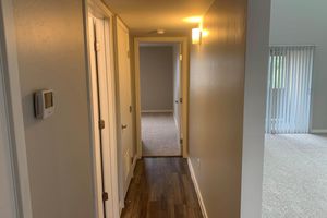A narrow hallway with light beige walls and wooden flooring. Two closed doors are visible on the left side, with a light fixture above. At the end of the hallway is a carpeted area leading to a room with vertical blinds on the right. The space appears well-lit and tidy.