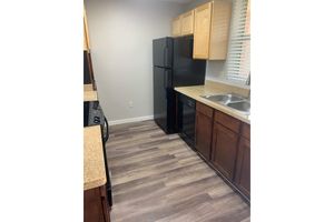 A modern kitchen featuring wooden cabinets, a black refrigerator, a black stove, and a double sink with a countertop. The flooring is a light wood laminate, and there are windows allowing natural light into the space. The walls are painted in a neutral color.