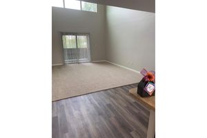 A spacious, empty room with light-colored walls and a large window letting in natural light. The floor features a mix of carpet and laminate. In the foreground, a gift wrapped with a bow sits on a kitchen counter. There are vertical blinds covering the window, and the overall ambiance is bright and airy.