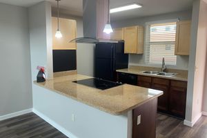 A modern kitchen featuring light wood cabinets, a black refrigerator, a sleek stove with an overhead vent, and a granite countertop. The space has recessed lighting and a window with blinds, creating a bright and inviting atmosphere. A small vase with flowers adds a touch of decor.