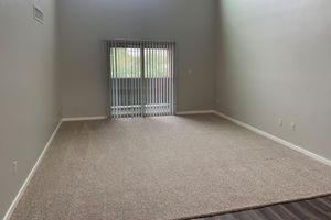 A spacious, empty room with light gray walls and plush beige carpet. There are large windows with vertical blinds allowing natural light to enter. The room has a high ceiling and a balcony door at one end, leading outside. The area is clean and well-maintained, suggesting a fresh or newly renovated space.