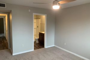 A well-lit room featuring carpeted flooring, a ceiling fan, and neutral-colored walls. The doorway leads to a bathroom area visible in the background, which includes a shower and toilet. The overall space appears clean and modern.