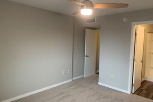 A well-lit, empty bedroom featuring beige walls and carpet flooring. A ceiling fan is visible, and an open white door leads to a bathroom area. The space is minimalist with no furniture, highlighting the room's dimensions and potential for personalization.