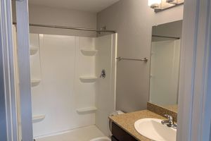 A clean, modern bathroom featuring a white shower stall with grab bars, a light brown granite countertop with a sink, a wall-mounted mirror, and beige walls. A towel rack is installed above the countertop, and a roll of toilet paper is visible on the side. Natural light illuminates the space from a wall-mounted light fixture.