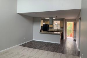 A spacious, well-lit open-concept living area featuring a carpeted space and modern kitchen with a countertop, light fixtures, and appliances. A sliding glass door leads to a small balcony with greenery visible outside, creating a bright and inviting atmosphere.