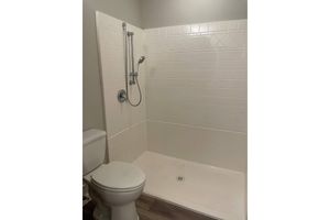 A clean, modern bathroom featuring a shower with a silver handheld showerhead affixed to a white tiled wall. The shower area is enclosed with white tiles, and a round drain is visible on the floor. To the right, a white toilet is positioned against the wall, complementing the restroom's simple design.