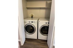 A side-by-side washing machine and dryer set in a small laundry space. They are both front-loading models with a simple white design, placed within a closet or designated laundry area. A shelf is above them, and there is a wall outlet visible.