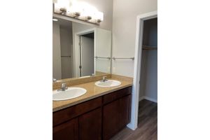 A modern bathroom featuring dual sinks with polished chrome faucets on a granite countertop. Above the sinks, there is a light fixture with several bulbs. A large mirror runs between the sinks. On the right, a doorway leads to a closet, and light-colored walls enhance the space's brightness.