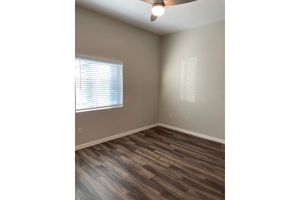 Empty room with light-colored walls and a hardwood floor. A window with white blinds allows natural light to enter, casting shadows on the wall. The ceiling features a ceiling fan. The overall atmosphere is bright and spacious, suitable for various uses like an office or bedroom.