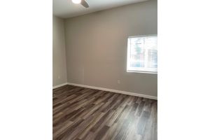 A partially furnished room with light gray walls and a window featuring white blinds. The floor is made of dark wood laminate, and a ceiling fan is visible. The room has no furniture, creating a spacious and empty feel.