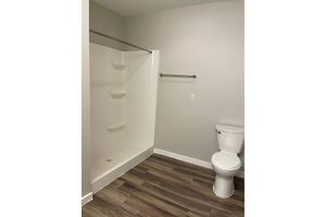 A clean, modern bathroom featuring a white shower stall with three built-in shelves, a chrome towel bar on the wall, and a white toilet. The floor is covered in dark wood laminate, and the walls are painted gray, creating a simple and contemporary aesthetic.