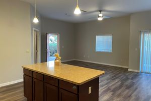 A spacious, modern kitchen and living area featuring a central island with a yellow vase, pendant lights hanging from the ceiling, light gray walls, and large windows. A door leads to an outdoor space, and there is neutral-colored flooring throughout the room.