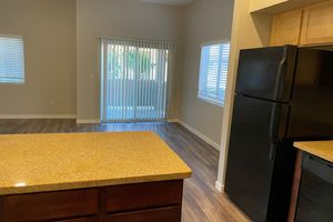 A spacious and modern kitchen and living area featuring a granite countertop island in the foreground, a black refrigerator, and light-colored cabinetry. The room has large windows with blinds, allowing natural light, and leads to a balcony. The flooring is a warm wood tone, enhancing the open and inviting atmosphere.