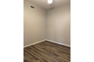A sparsely furnished room with beige walls and a light-colored wooden floor. There is a ceiling fan visible in the upper corner, and the room features an empty space with no furniture, creating a minimalist appearance. Natural light may enter through a nearby window not shown in the image.