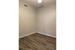 A well-lit, empty room with light gray walls and a ceiling fan. The floor is covered with wood-like laminate flooring. There are no furnishings or decorations, creating a spacious and minimalistic feel. The room features two electrical outlets on the walls.
