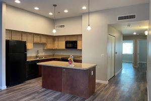Modern kitchen featuring wooden cabinetry, granite countertops, and stainless steel appliances, including a black refrigerator and microwave. Two pendant lights hang above the kitchen island. Light-colored walls and laminate flooring create a bright atmosphere, with a doorway leading to a hallway in the background.