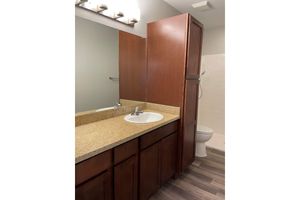 A small bathroom featuring a wooden vanity with a sink and a countertop. A mirror is mounted above the sink, and there is a tall cabinet beside it. The space includes a white toilet and a shower area with white tiles. The flooring is a light-colored laminate, creating a modern and tidy atmosphere.