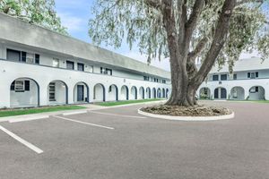 A courtyard view of a two-story, white building featuring arched doorways. In the center, a large tree adds greenery, surrounded by a circular driveway. The scene is bright and inviting, with a clear blue sky overhead and parking spaces nearby.