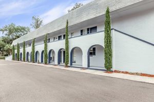 A light blue building featuring multiple arches along the facade, flanked by tall, slender green shrubs. The structure has a sloping gray roof and is set on a quiet street with a clear sky in the background. The overall design reflects a modern aesthetic with a tranquil atmosphere.