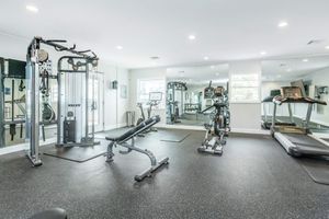 A bright and spacious modern gym featuring various exercise equipment including a cable machine, weight bench, stationary bike, and treadmill. The room has mirrors on one wall and is equipped with overhead lighting, creating an inviting atmosphere for workouts.