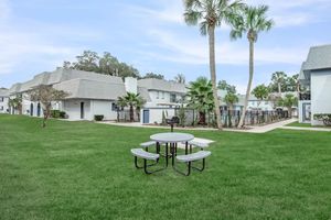 A tranquil outdoor area featuring a picnic table with benches, surrounded by well-maintained grass and palm trees. In the background, there are residential buildings and a fenced swimming pool area, ideal for relaxation and leisure activities.