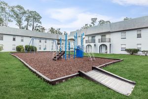 A children's playground with blue equipment, including slides and swings, situated in a grassy area surrounded by mulched ground. The playground is located in the courtyard of a white apartment building with balconies. Trees are visible in the background under a clear sky.
