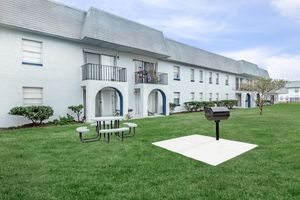 A grassy area with a picnic table and grill in front of a light blue, multi-unit residential building. The building features arched doorways, balconies, and windows, surrounded by manicured shrubs. The sky is clear and bright, creating a welcoming outdoor space.