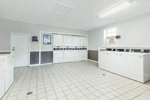 A clean, well-lit laundry room featuring multiple white washing machines and dryers along one wall. There are tiled floors, a door with a small window, and a wall-mounted dispenser. The room has neutral-colored walls and large windows allowing natural light.