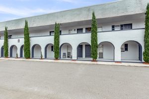 A low-rise building featuring a series of arches along its facade, with two tall, narrow green trees flanking the entrance. The exterior is painted light gray, and there are dark blue doors and windows. A small chair is visible on the porch. The area in front is paved and lined with landscaping.