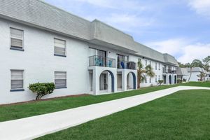 A landscaped pathway leads past a two-story apartment building with a white exterior and arched doorways. Green grass surrounds the building, and blue chairs are visible on balconies. The sky is partly cloudy, contributing to a serene residential atmosphere.