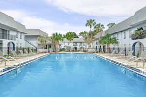 A serene view of a swimming pool surrounded by lounge chairs in a well-maintained courtyard. Palm trees and greenery add to the tropical atmosphere. The buildings in the background are light-colored with balconies, creating a relaxing ambiance, perfect for leisure and relaxation.
