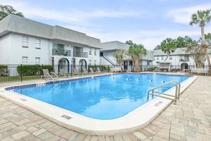 A clear blue swimming pool surrounded by lounge chairs, set in a landscaped area with palm trees. In the background, there are two-story light-colored apartment buildings with balconies. The scene is sunny and inviting, creating a relaxing atmosphere.