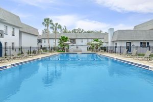 A clear and inviting swimming pool surrounded by lounge chairs, set in a residential area with palm trees and light-colored apartment buildings in the background. The sky is blue with a few clouds, creating a serene atmosphere for relaxation and leisure.