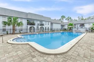 A clear blue swimming pool surrounded by paved decking, with palm trees and green plants nearby. The pool is located in a residential area with light gray buildings in the background and balconies visible. The sky is bright with a few clouds, creating a relaxing atmosphere.