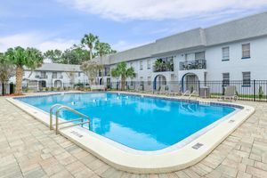 A clear blue swimming pool surrounded by a spacious patio area, with several lounge chairs and palm trees nearby. The pool is situated in front of a light-colored, two-story building with balconies. The setting is peaceful and inviting, ideal for relaxation.