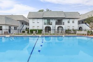 A clear blue swimming pool surrounded by lounge chairs and a safety fence, with two-story apartment buildings in the background. The sky is partly cloudy, adding to a relaxing atmosphere.