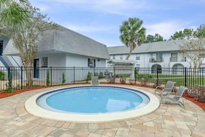 A small round swimming pool surrounded by a stone patio and a black fence. In the background, there are gray buildings with a sloped roof and green landscaping, including palm trees and shrubs. Two deck chairs are placed beside the pool, creating a relaxing outdoor space.