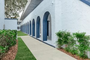 A pathway lined with neatly trimmed grass and shrubs leads to a series of arched doorways on a white building. The architecture features blue accents and a smooth concrete walkway, creating a clean and inviting atmosphere. The scene is well-lit, showcasing the building's exterior details.