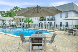 A serene swimming pool surrounded by lounge chairs and tables, with a large umbrella providing shade. The area is landscaped with palm trees, and adjacent buildings feature multiple balconies. A relaxing outdoor space perfect for leisure and socializing.