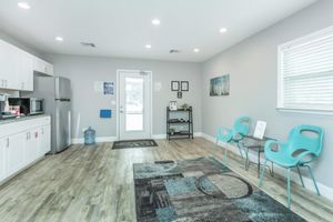 Bright and airy waiting area with light gray walls and wooden flooring. Features include a kitchenette with a refrigerator, a water cooler, and seating with modern turquoise chairs. A small shelf with decorative items and a door leading outside are visible. A rug with geometric patterns adds warmth to the space.