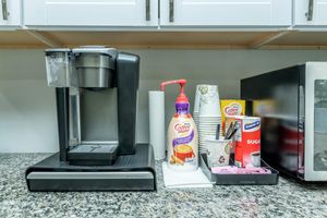 A coffee maker sits on a granite countertop alongside a container of sugar, Coffee-Mate creamer, cups, and a pump dispenser. There’s also a microwave in the background. The setting appears to be a kitchen with white cabinets above.
