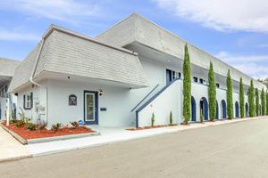A light blue building with a modern design featuring a triangular roof and several arched windows. It has a sign indicating the name of the building and is surrounded by neatly maintained landscaping, including small shrubs and decorative gravel. The scene is set on a sunny day with a clear sky.