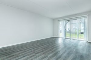 A spacious, empty room with light gray walls and a wooden floor. Large sliding glass doors with vertical blinds lead to an outdoor area, allowing natural light to fill the space. The room is unfurnished, creating a minimalist and open atmosphere.