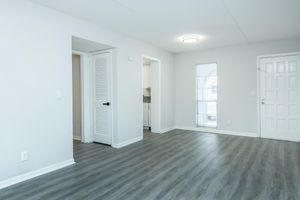 A bright, modern living space featuring light gray walls and new laminate flooring. The room includes a door on the right leading outside, a window with blinds for natural light, and a partially visible doorway leading to another room. The ceiling is simple, with a modern light fixture.