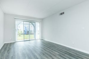 A light and airy empty room with gray laminate flooring, featuring white walls and a sliding glass door with vertical blinds. The door leads to an outdoor area, and there's natural light entering the space, creating an inviting atmosphere. A small area for potential furniture placement is visible.