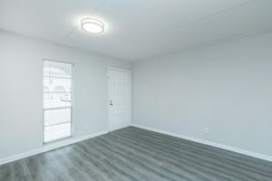 A bright, empty room with a light fixture on the ceiling. The walls are painted light gray, and there is a door on one side. A window with blinds allows natural light to enter, illuminating the light-colored flooring. The overall atmosphere is clean and minimalistic.