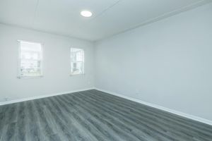 A bright, empty room with a light-colored wall and large windows featuring white blinds. The floor has a sleek, gray laminate finish. The ceiling is plain with a circular light fixture. The space is minimalistic, creating a clean and open atmosphere.