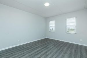 A light and airy empty room with gray laminate flooring. There are two windows with white blinds, allowing natural light to enter. The walls are painted in a light color, creating a spacious feel. The ceiling has a simple round light fixture. No furniture or decorations are present.