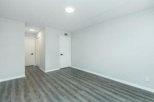 A spacious, empty room with light gray walls and a wood-like floor. There is a closed door on the right leading to another room and a doorway on the left, leading to a hallway. Ceiling lights provide bright illumination. The room is devoid of furniture, creating an open, minimalist feel.